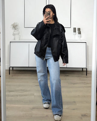 Femme portant une veste bomber oversize noire effet cuir, avec un jean bleu clair, prenant une photo dans un intérieur minimaliste.