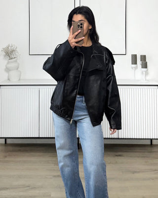 Femme portant une veste bomber oversize noire effet cuir, avec un jean bleu clair, prenant une photo dans un intérieur minimaliste.