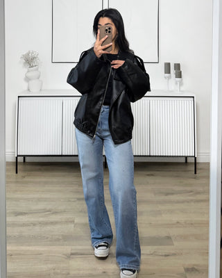 Femme portant une veste bomber oversize noire effet cuir, avec un jean bleu clair, prenant une photo dans un intérieur minimaliste.
